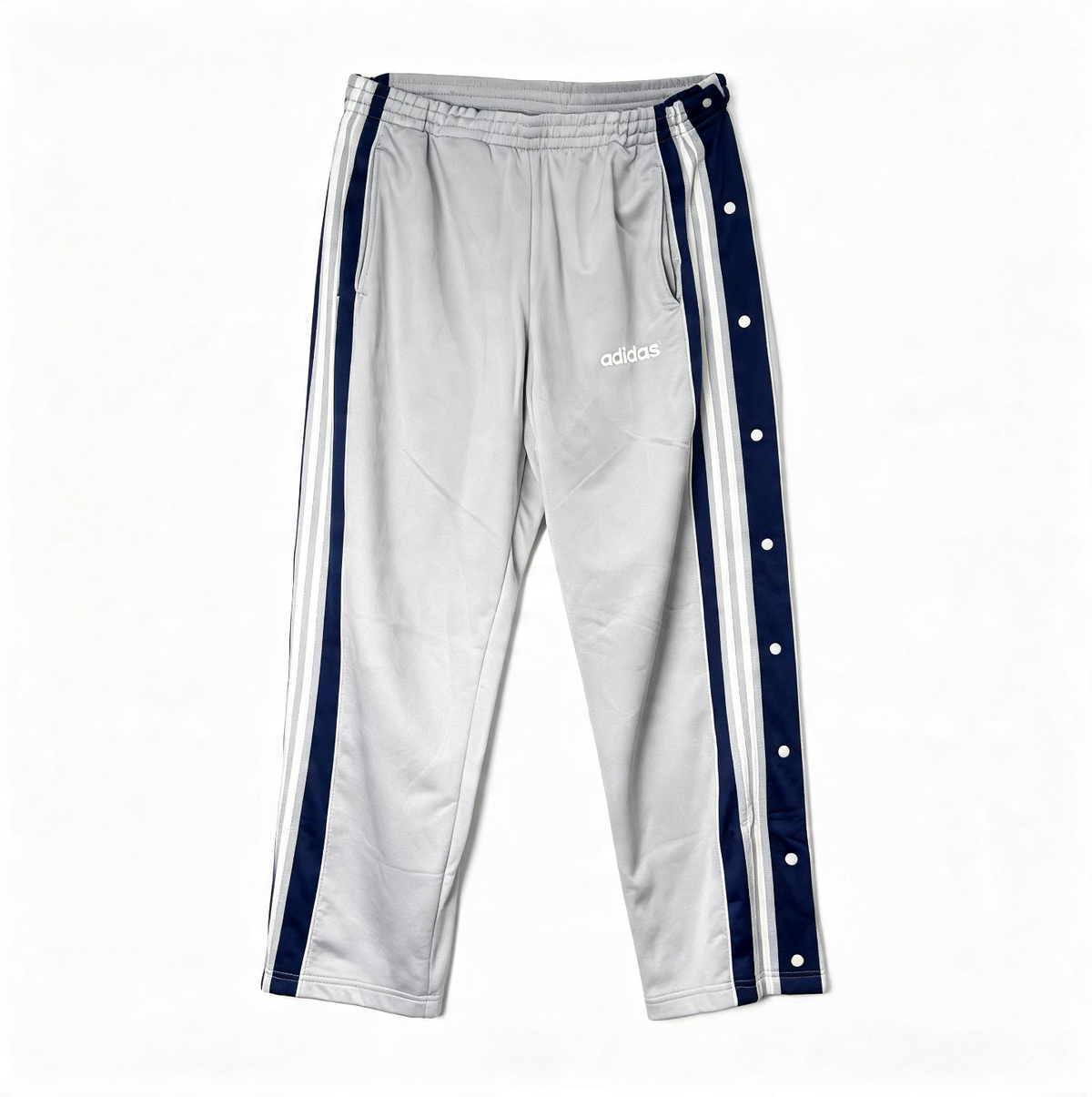 A pair of Adidas track pants in light gray, featuring navy blue side stripes with white snap buttons and a white Adidas logo on the left thigh, laid flat against a white background.