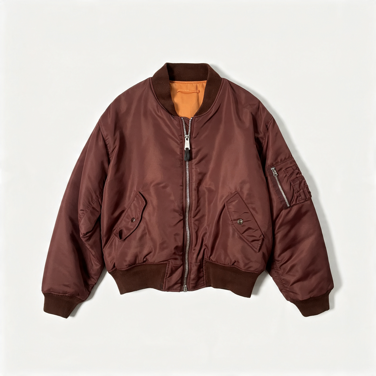 A classic bomber jacket in a deep reddish-brown color, featuring a full front zipper, ribbed knit collar, cuffs, and hem, with zippered utility pockets on the chest and sleeve, laid flat against a light off-white background.