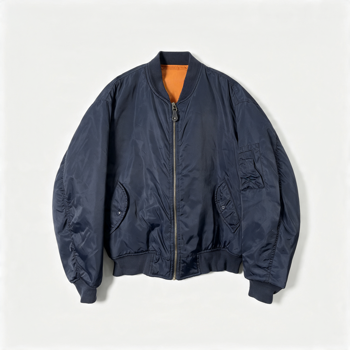 A navy blue nylon bomber jacket with a gold-tone zipper, ribbed collar, cuffs, and hem, featuring a bright orange interior lining, laid flat against a light gray background.