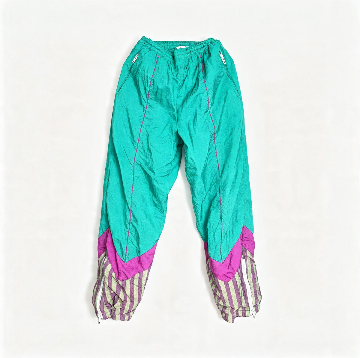 A pair of vintage windbreaker pants in a vibrant turquoise green, featuring magenta pink chevron details at the lower legs and striped purple-and-white cuffs, laid flat against a white background.