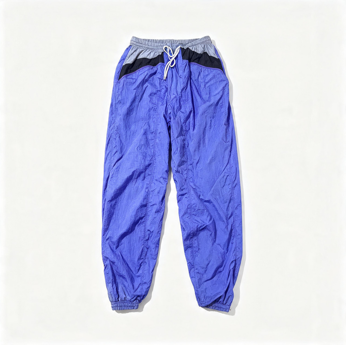 A pair of loose-fit jogger-style pants laid flat on a light off-white background. The pants are primarily a vibrant periwinkle blue with a contrasting light gray waistband, a black diagonal stripe across the hips, and a white drawstring.