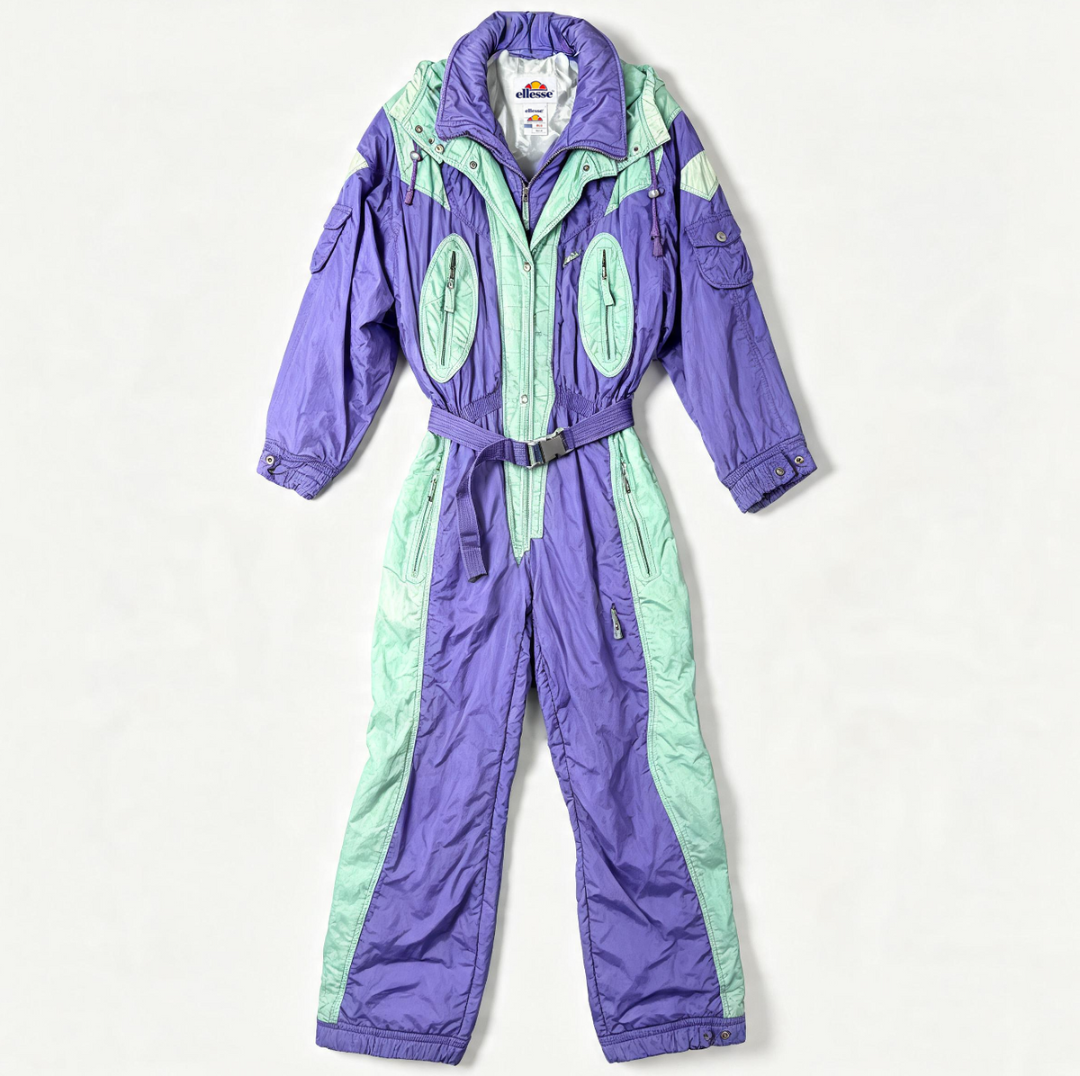 A vintage snowsuit in purple and mint green, laid flat against a light off-white background. The one-piece jumpsuit features a purple collar, purple legs, and mint green side panels and torso accents.