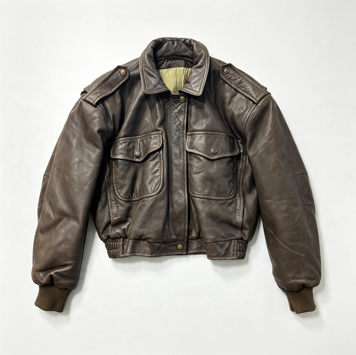 A brown leather jacket with shoulder epaulets, flap chest pockets, and ribbed cuffs and hem, laid flat against a light gray background.