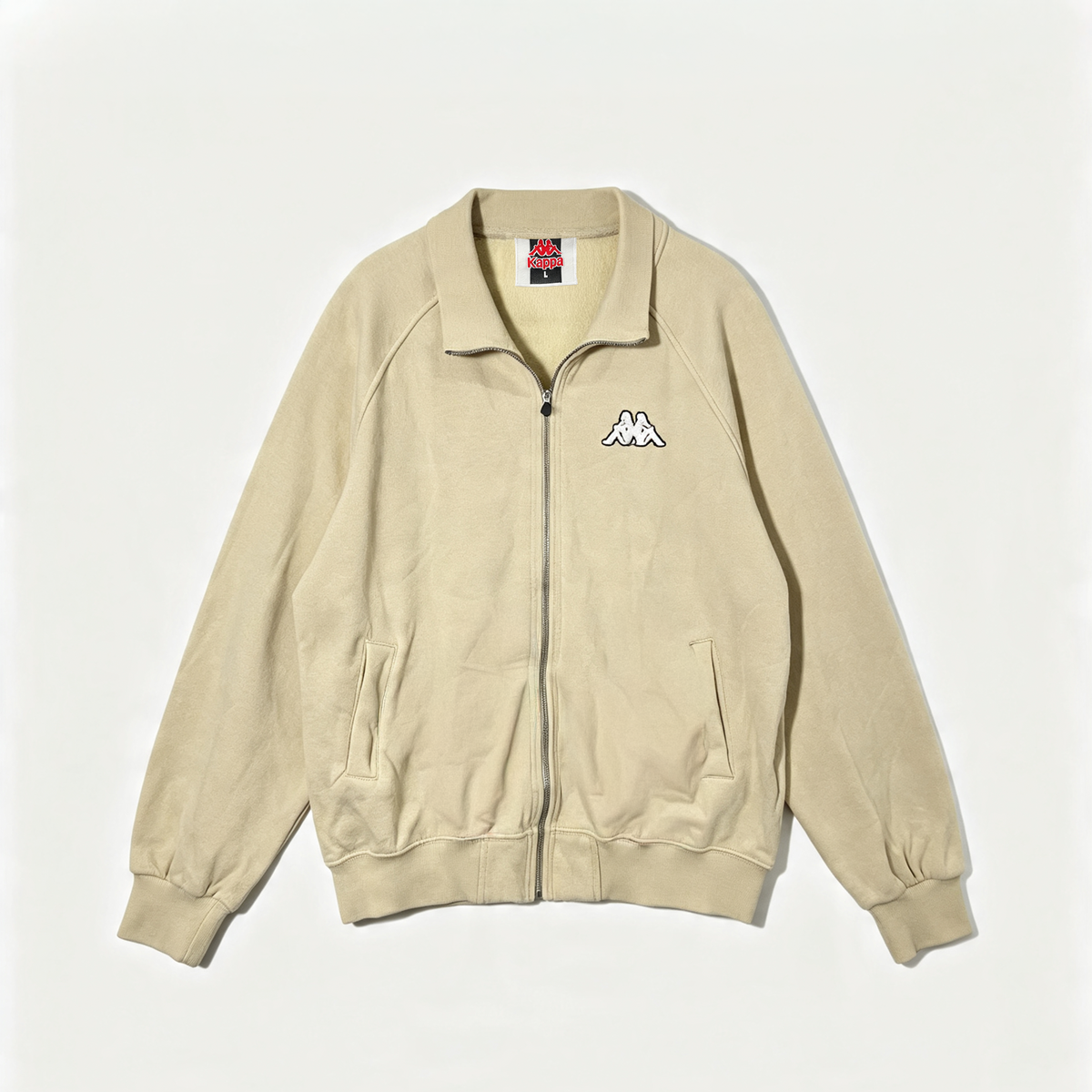 A beige zip-up jacket with a stand collar, featuring the Kappa double-M logo embroidered in white on the left chest, laid flat against a light off-white background.