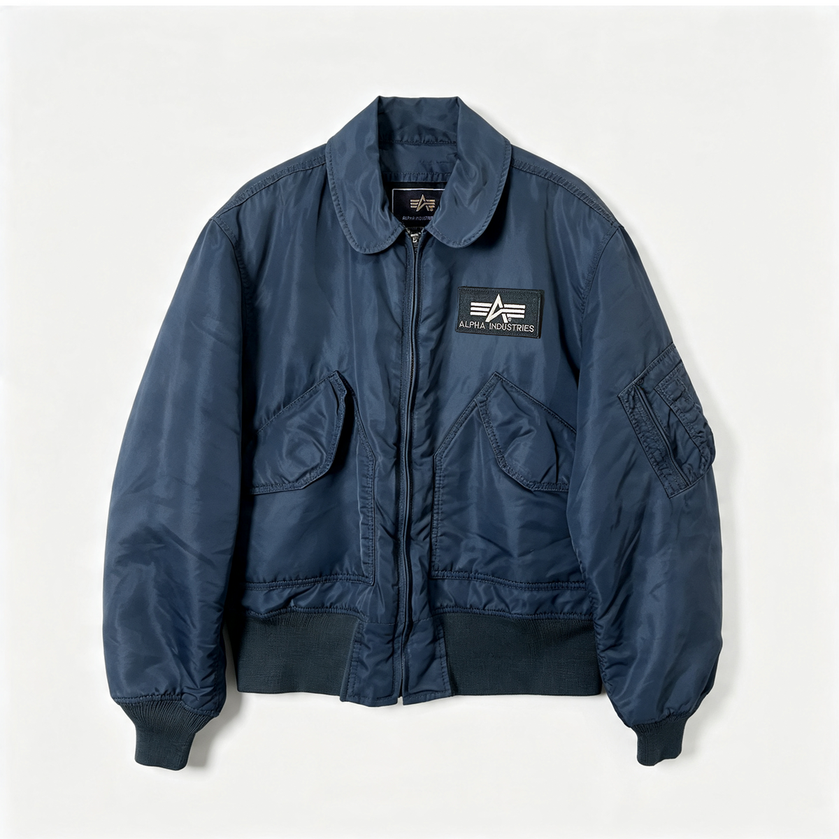 A navy blue Alpha Industries bomber jacket with a unique rounded collar, front zipper, and two large front flap pockets, laid flat against a light off-white background.