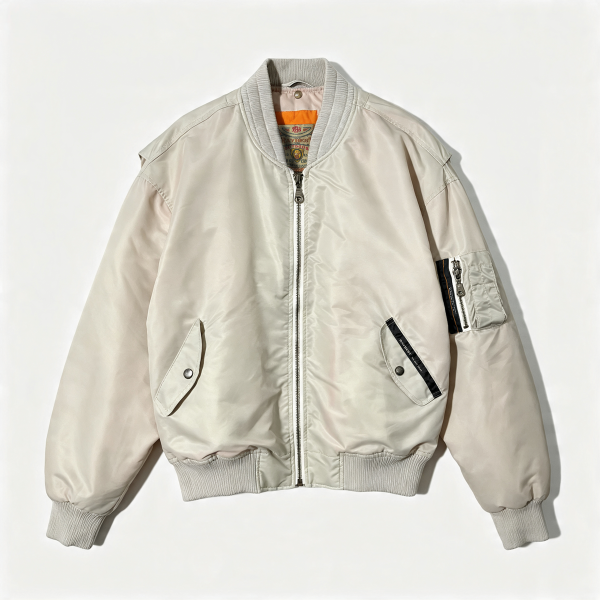A light beige nylon bomber jacket with ribbed collar, cuffs, and hem, featuring a front zipper, diagonal welt pockets, and a zippered sleeve patch pocket, shown flat on a light background.