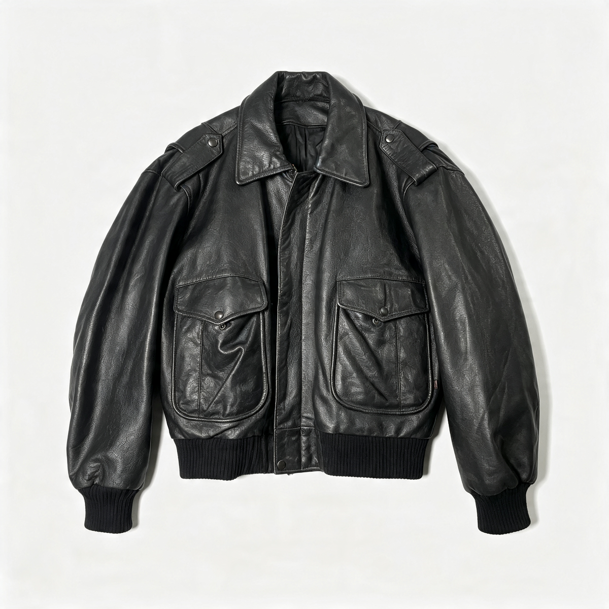 A black leather bomber jacket with a pointed collar, shoulder epaulets, two large front flap pockets, and ribbed knit cuffs and hem, laid flat against a light off-white background.