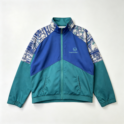 A colorblock zip-up jacket with a royal blue yoke, teal lower body and sleeves, and a blue-and-white graphic pattern on the upper sleeves, shown flat against a light gray background.