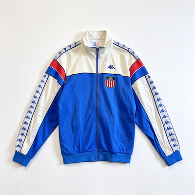 A vintage Kappa track jacket in royal blue, white, and red, featuring a crest on the left chest and Kappa logo on the right chest. The sleeves have blue Kappa logos printed down the white panels.