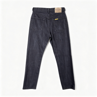 Back view of dark washed denim jeans with a tan leather brand patch on the waistband and a small yellow accent tab on the back pocket, laid flat on a white background.