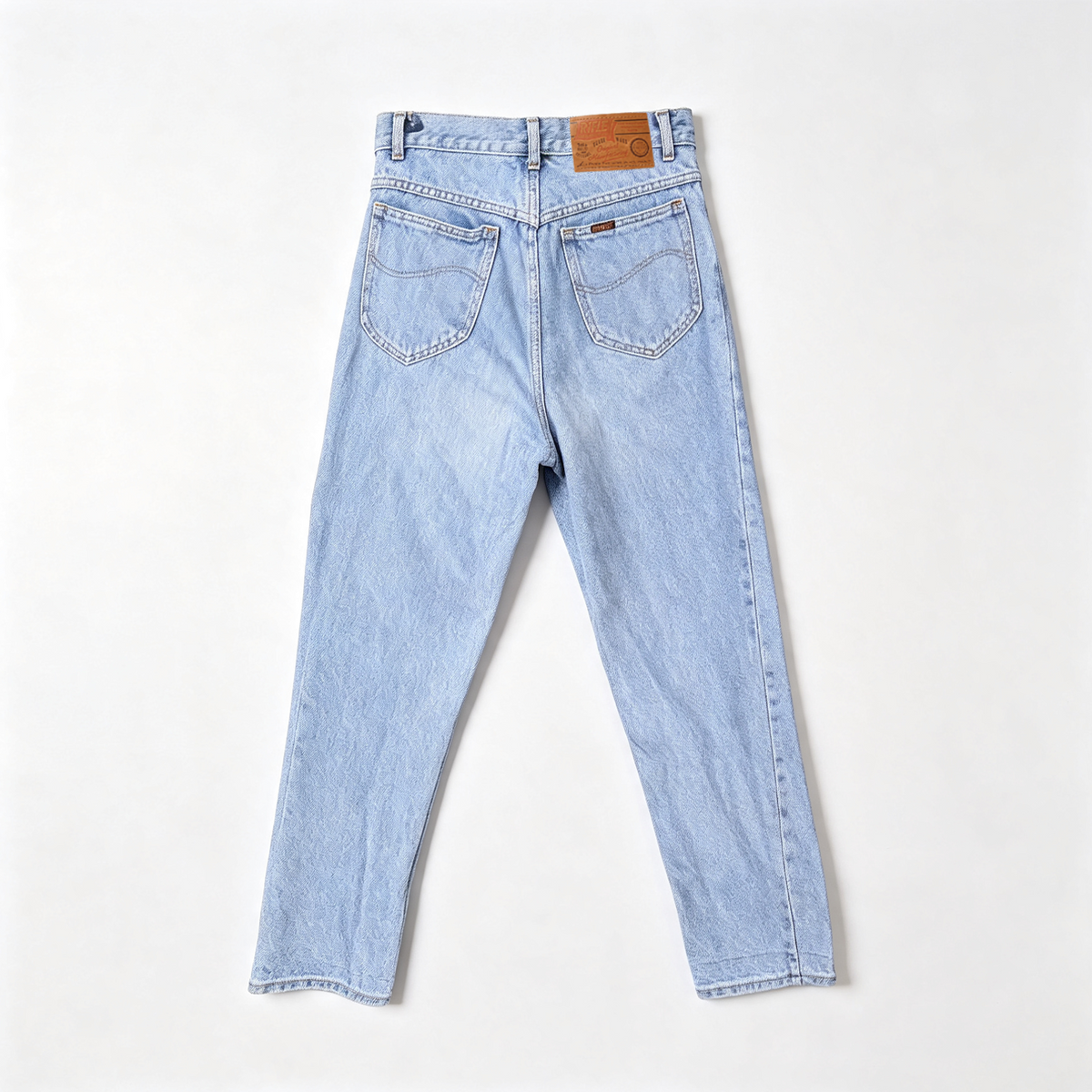 Light wash blue denim jeans shown from the back, featuring a classic five-pocket design and a brown leather patch on the back waistband, laid flat against a light gray background.
