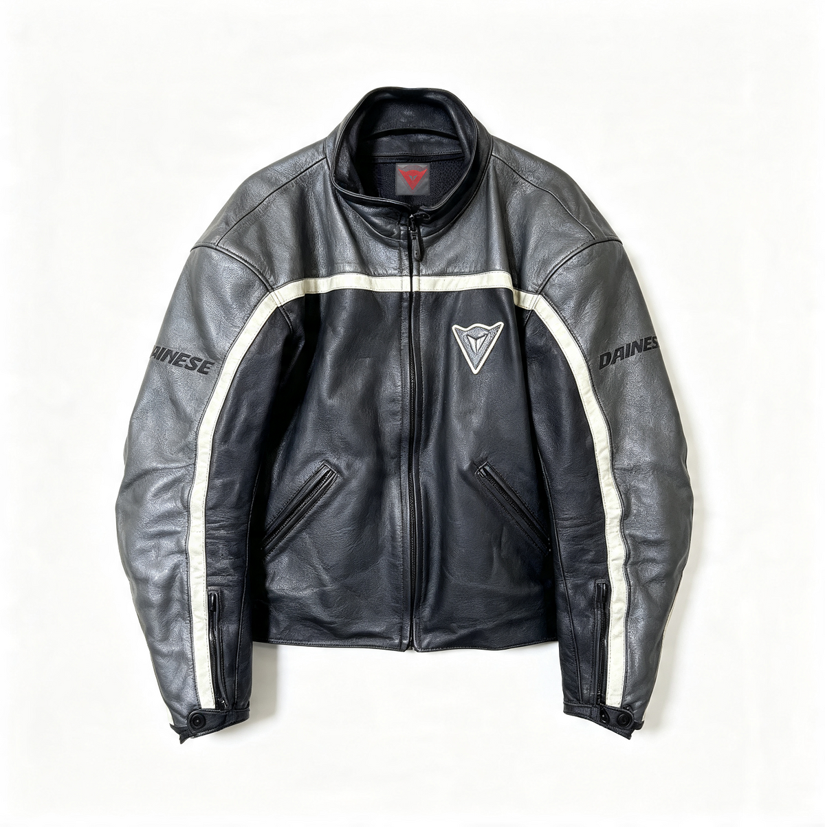 A black leather motorcycle jacket with white stripe details on the sleeves, featuring the Dainese brand name and logo, laid flat against a white background.