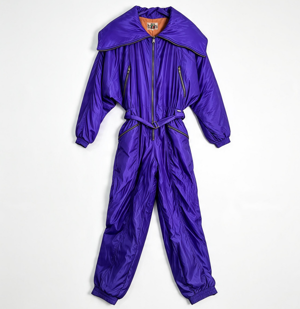 A shiny purple one-piece puffer jumpsuit with a large collar, front zipper, zip pockets, and a matching waist tie, laid flat on a white background.