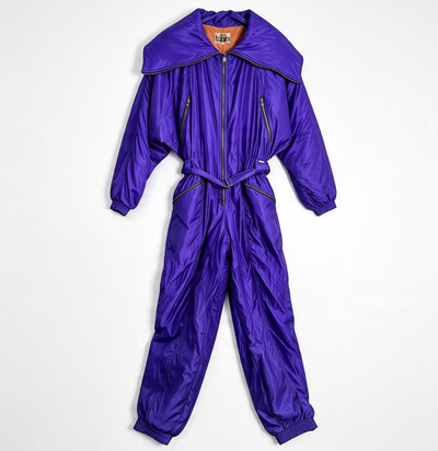 A shiny purple one-piece puffer jumpsuit with a large collar, front zipper, zip pockets, and a matching waist tie, laid flat on a white background.