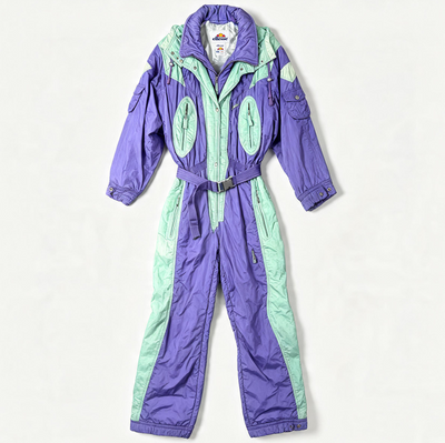 A vintage snowsuit in purple and mint green, laid flat against a light off-white background. The one-piece jumpsuit features a purple collar, purple legs, and mint green side panels and torso accents.