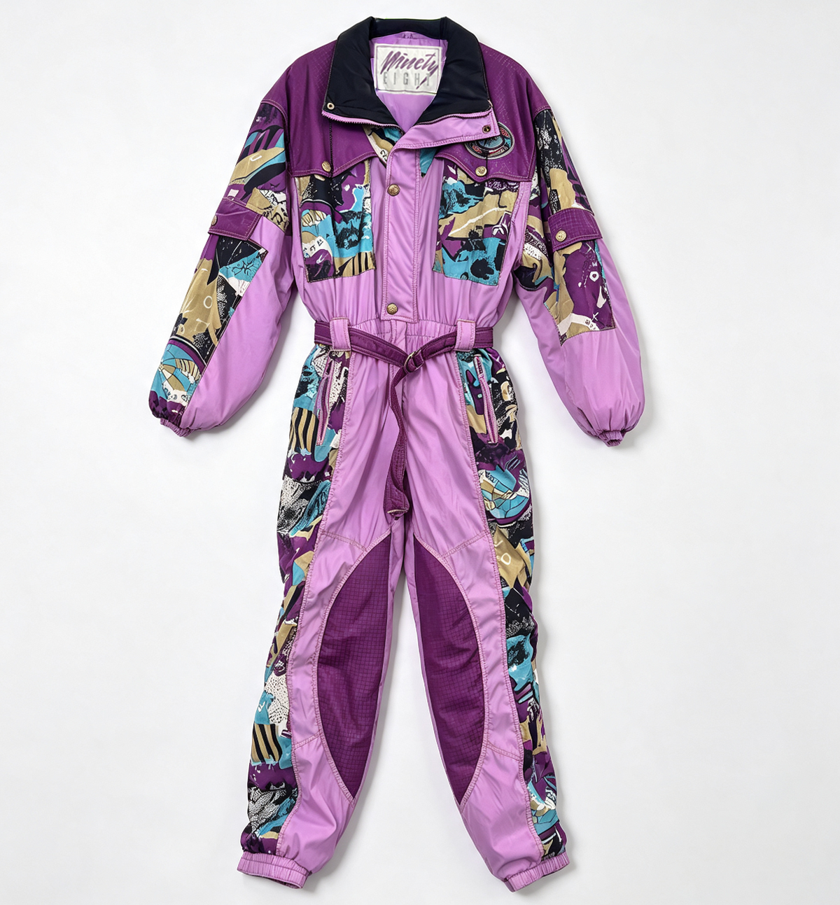 A vintage one-piece jumpsuit in purple, featuring a multicolor graphic print with cartoon characters and panels of solid purple. The jumpsuit has a zippered chest, snap buttons, a matching purple waist belt, and a black collar.