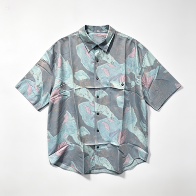 A short-sleeve button-up shirt with a tropical leaf pattern in shades of gray, teal, and pink, laid flat against a light gray background.